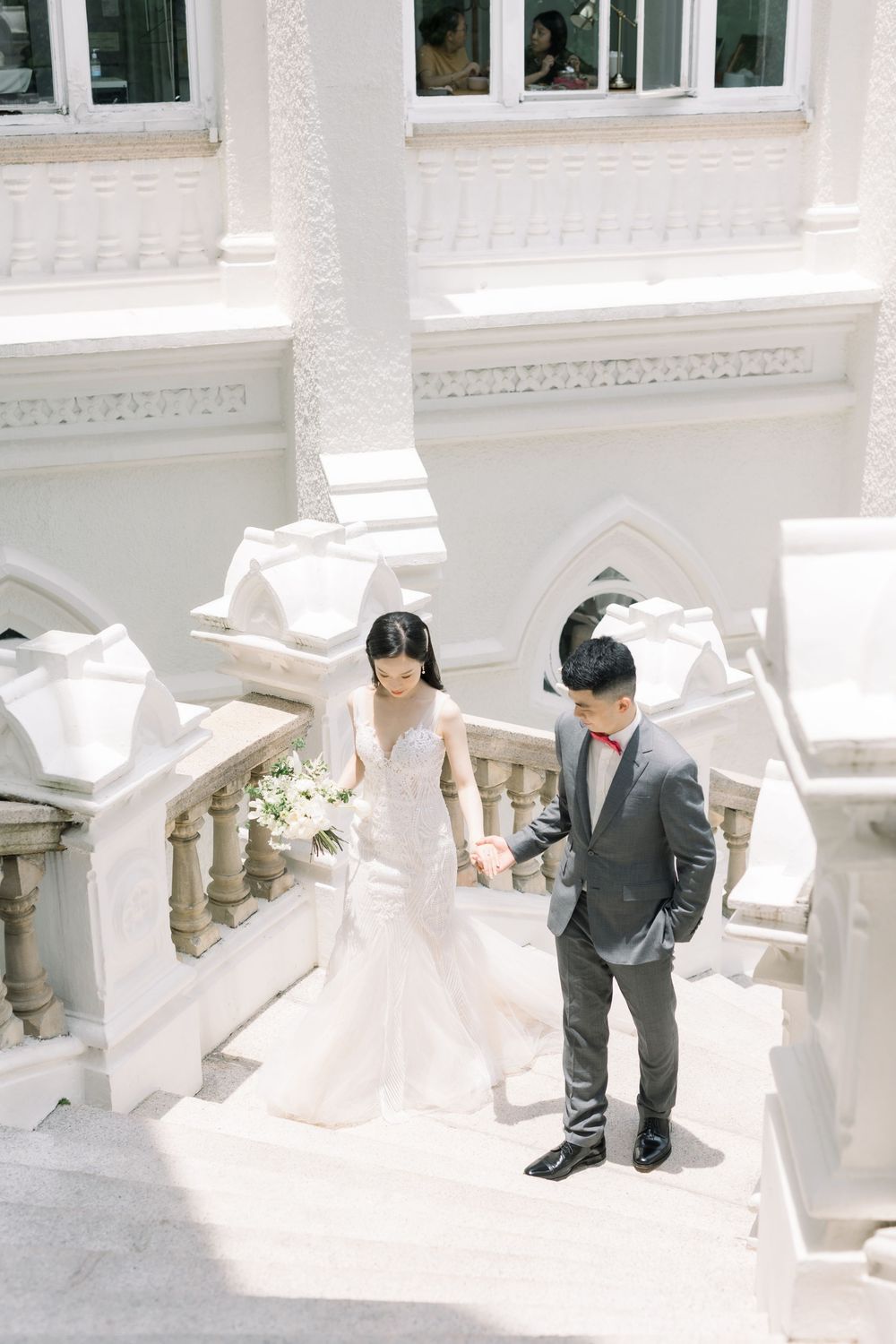 A couple in wedding attire poses on white stone steps in an elegant architectural setting with natural lighting.