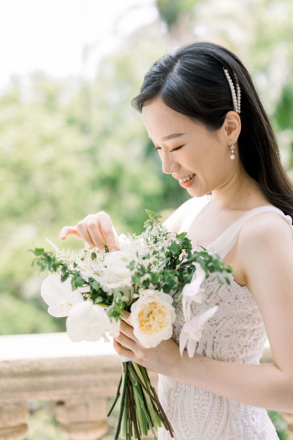 A bride in a lace dress smiles while holding a romantic bouquet of blush and cream roses with greenery.