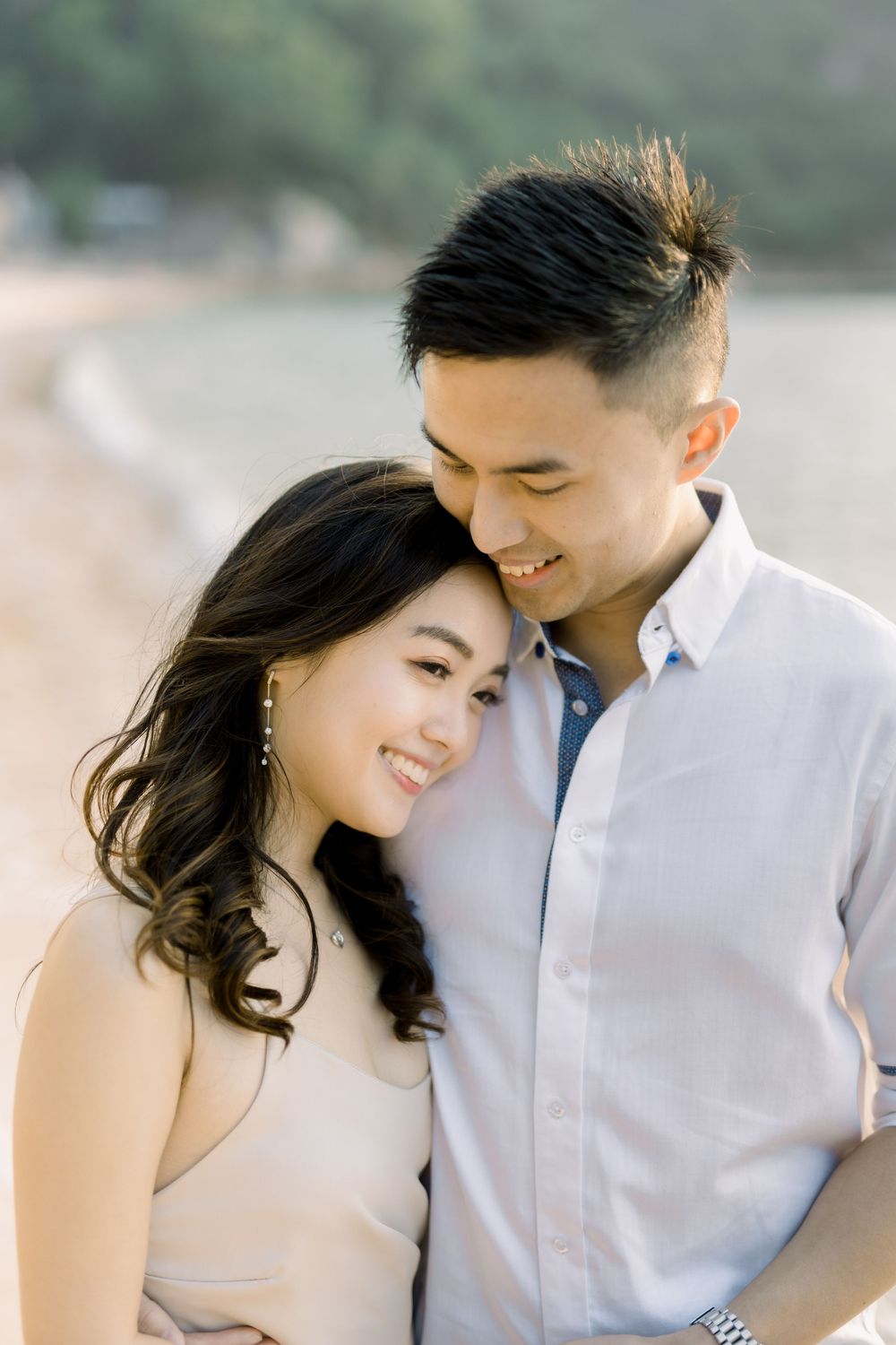 A romantic couple in white attire shares intimate moments during their engagement photoshoot by the water.