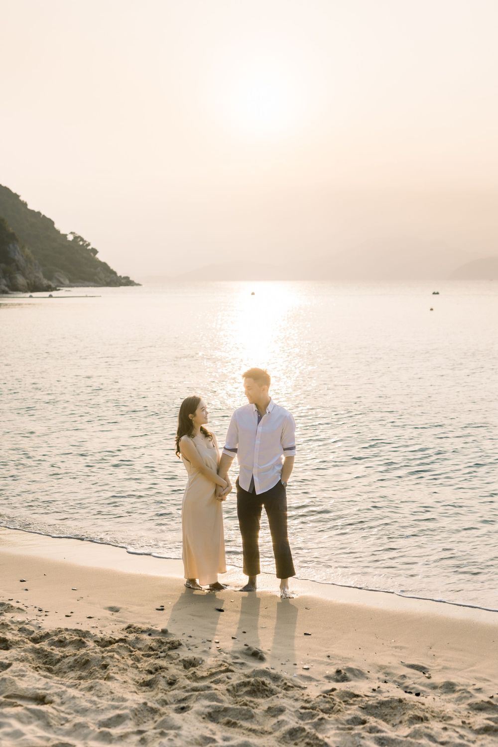 A romantic couple stands together on a beach at sunset with mountains and ocean in the background.