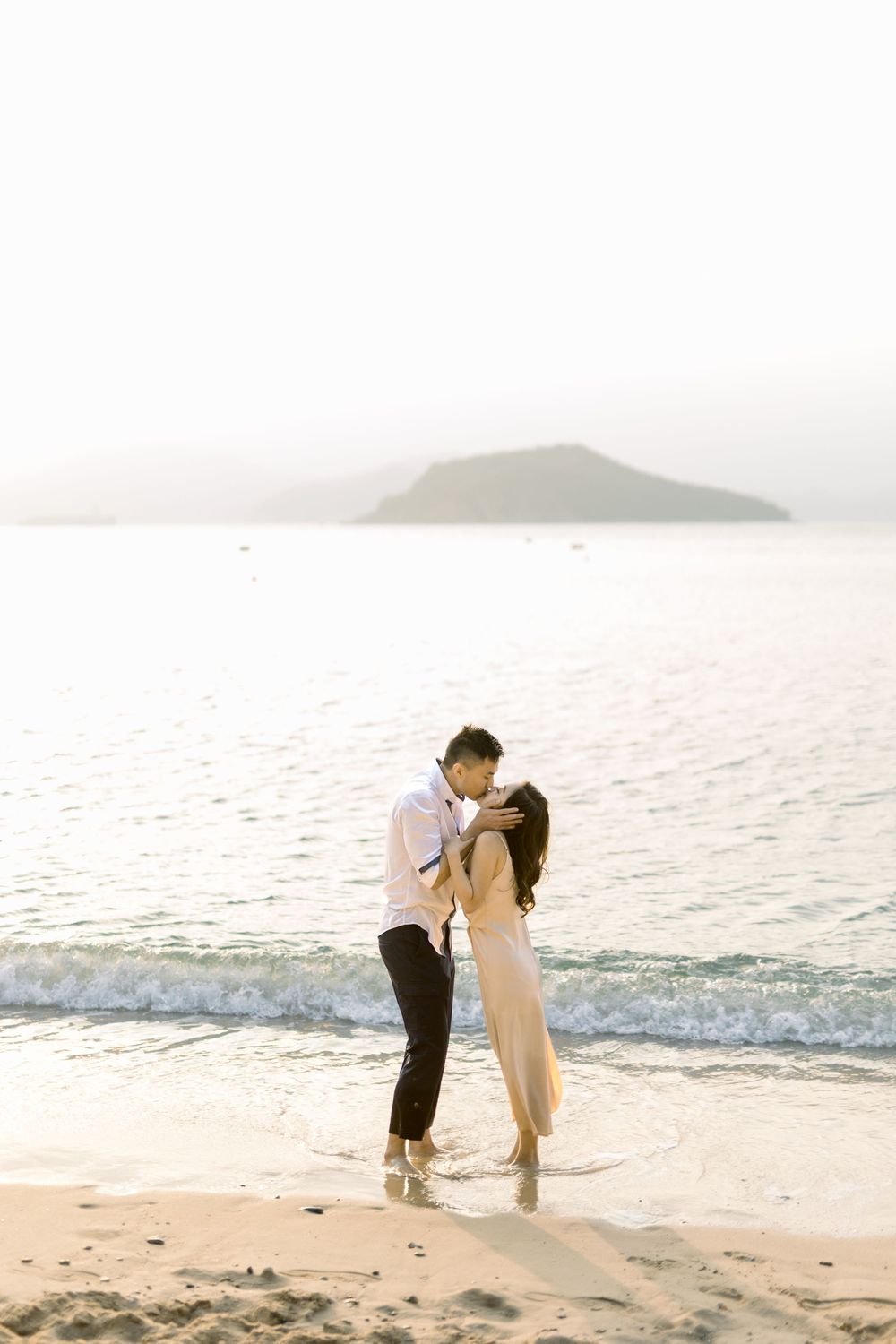 A couple embraces on the beach with mountains and ocean views at sunset.