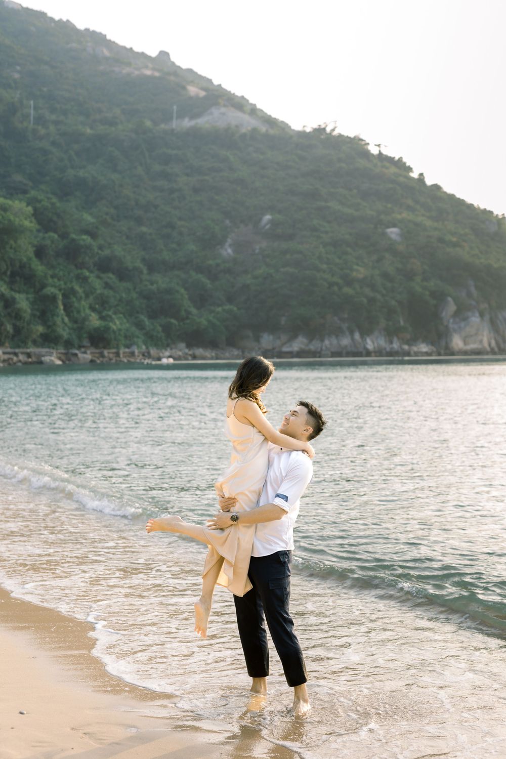 A couple dances playfully in the shallow water at a scenic beach with mountains in the background during sunset.