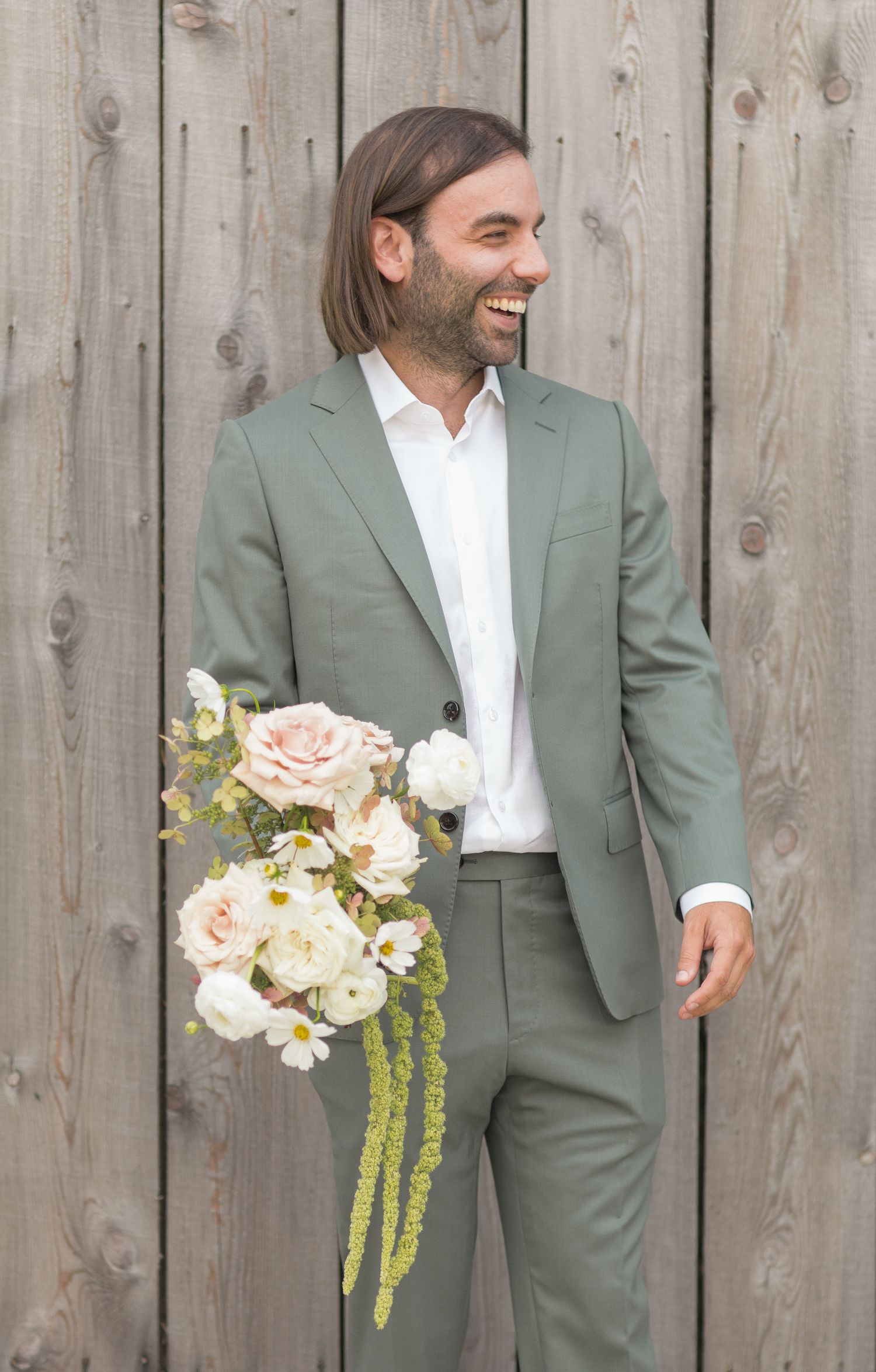 A person in a sage green suit and white shirt laughs joyfully while holding a bouquet of blush and cream flowers.
