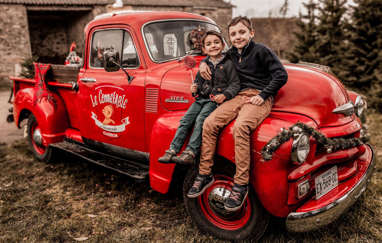 A classic red vintage Chevrolet pickup truck with two people sitting on its fender in a rustic outdoor setting.