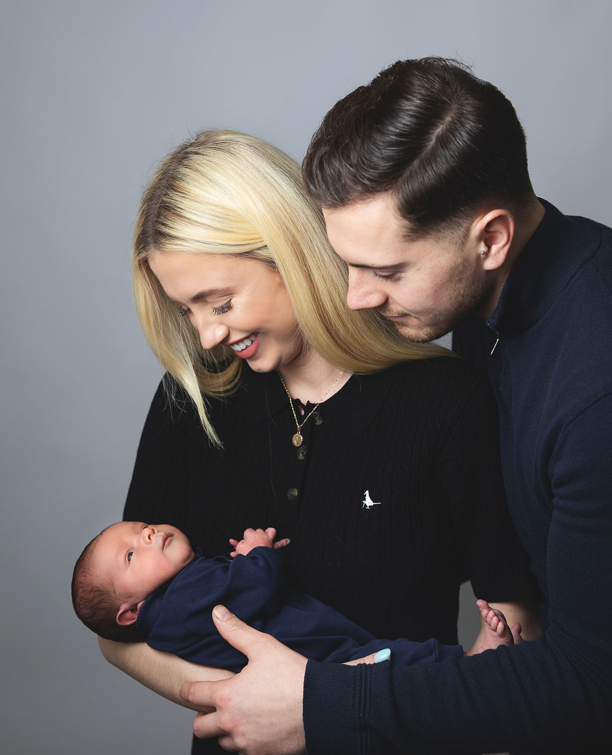 Parents sharing a joyful moment with their newborn baby wearing blue clothing against a grey backdrop. During a newborn session in Sidcup