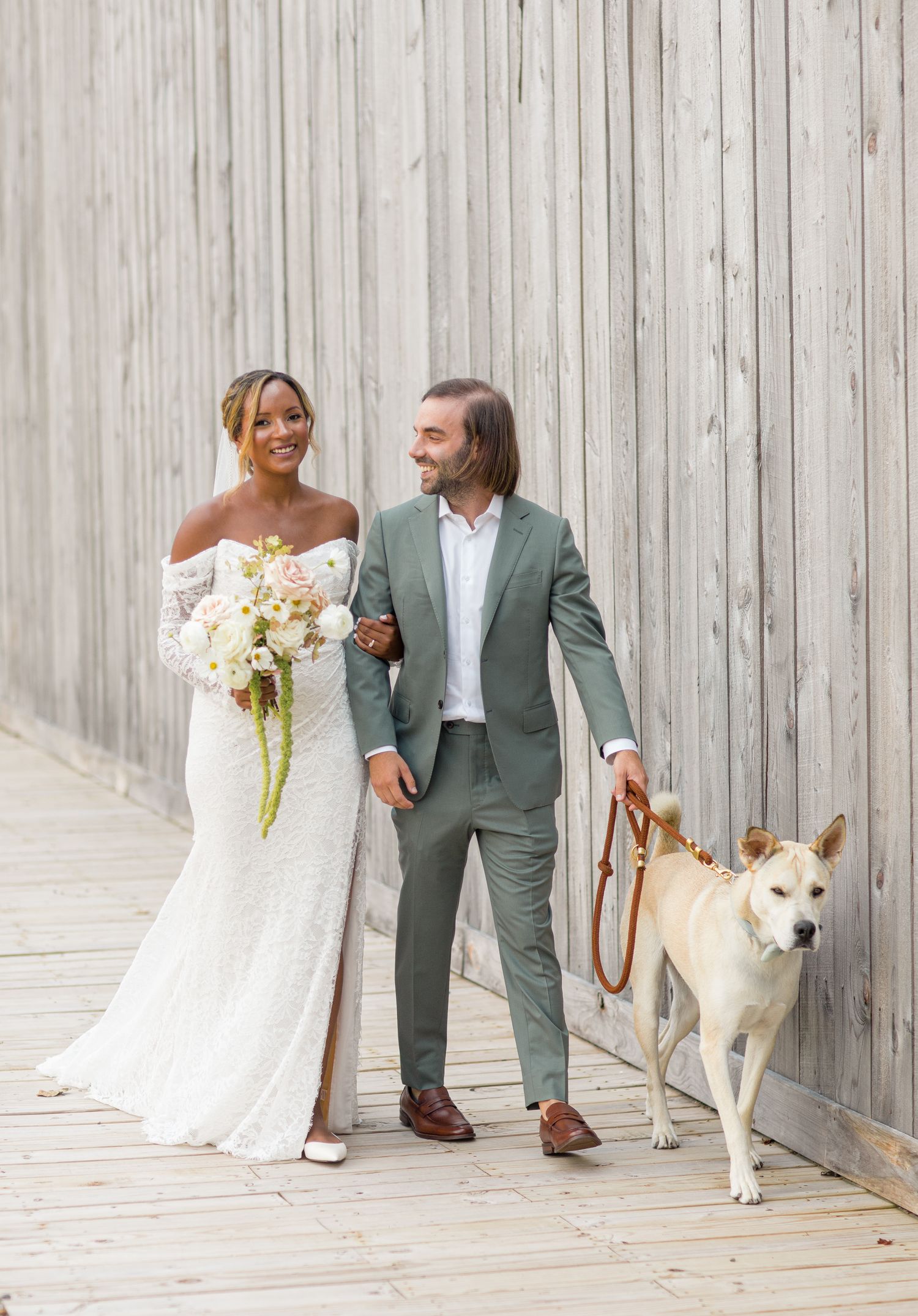 Happy couple in wedding attire poses together on a wooden deck with their white dog on a rustic background.