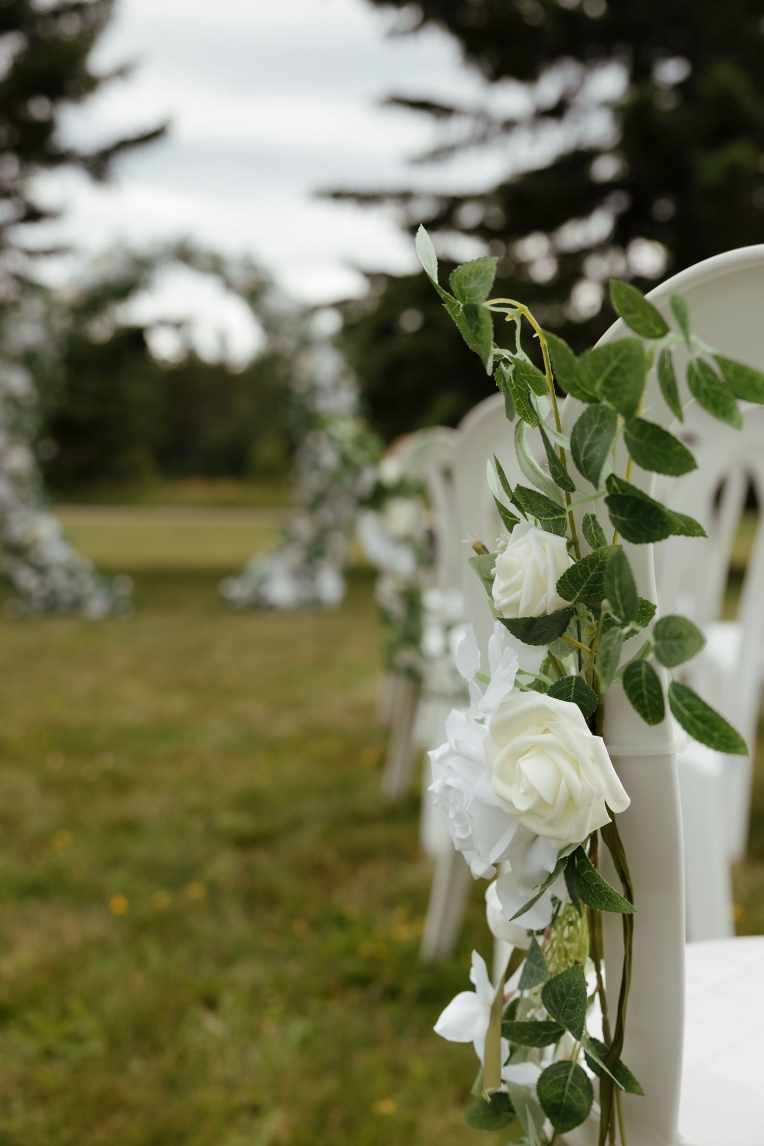 White wooden posts decorated with elegant white roses and green foliage create an outdoor wedding aisle setting.