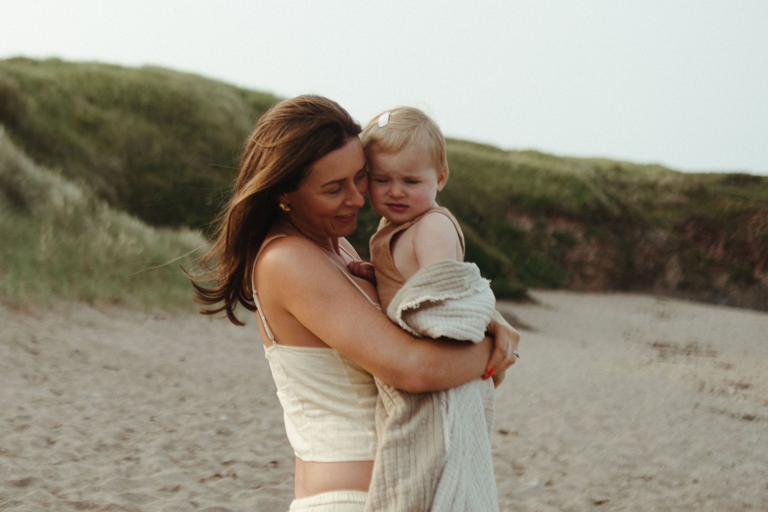 Mother and baby share tender moments during a beach photography session on sandy coastal dunes at sunset.