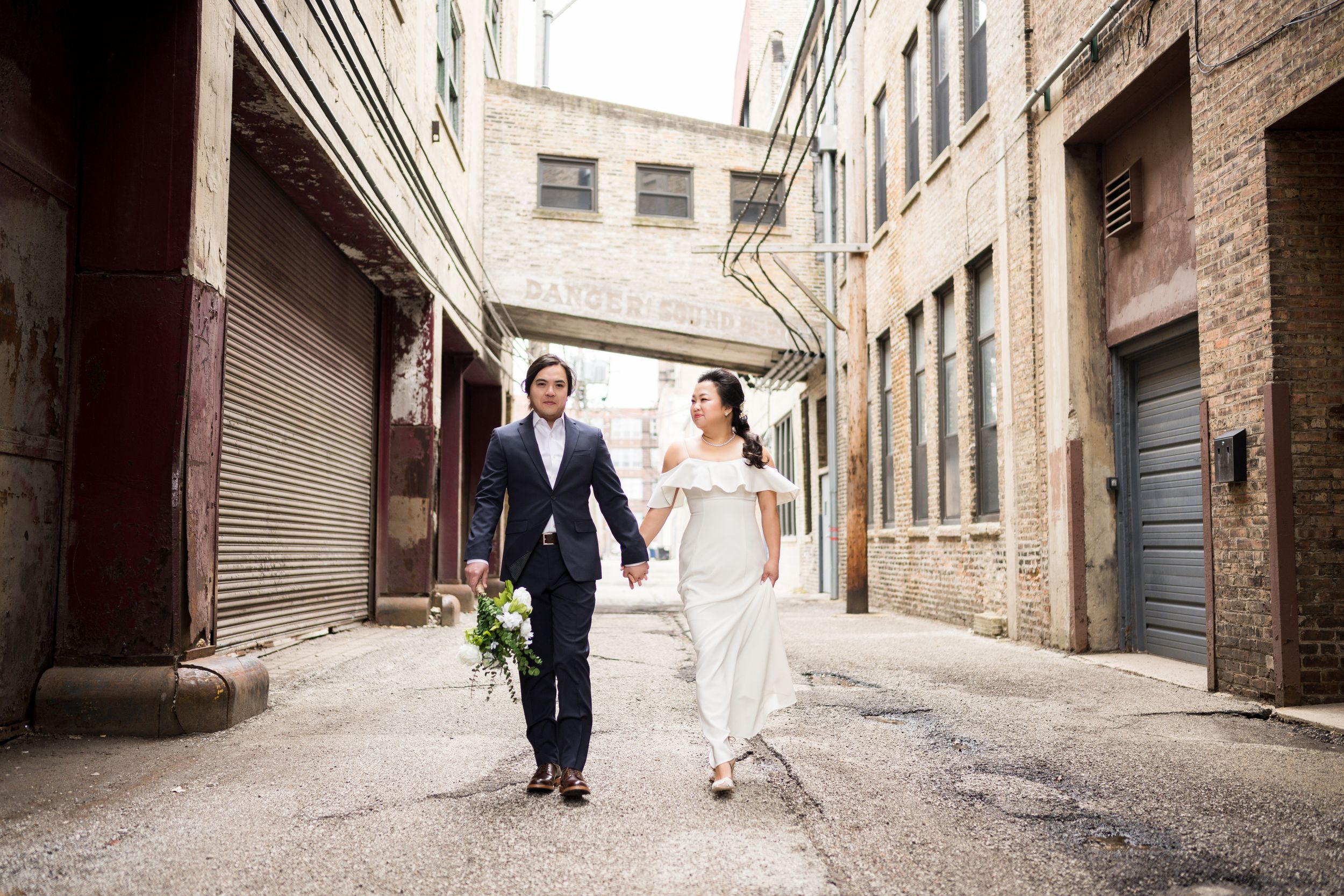 A couple in wedding attire walks through a rustic alleyway with brick walls and cobblestone paving.