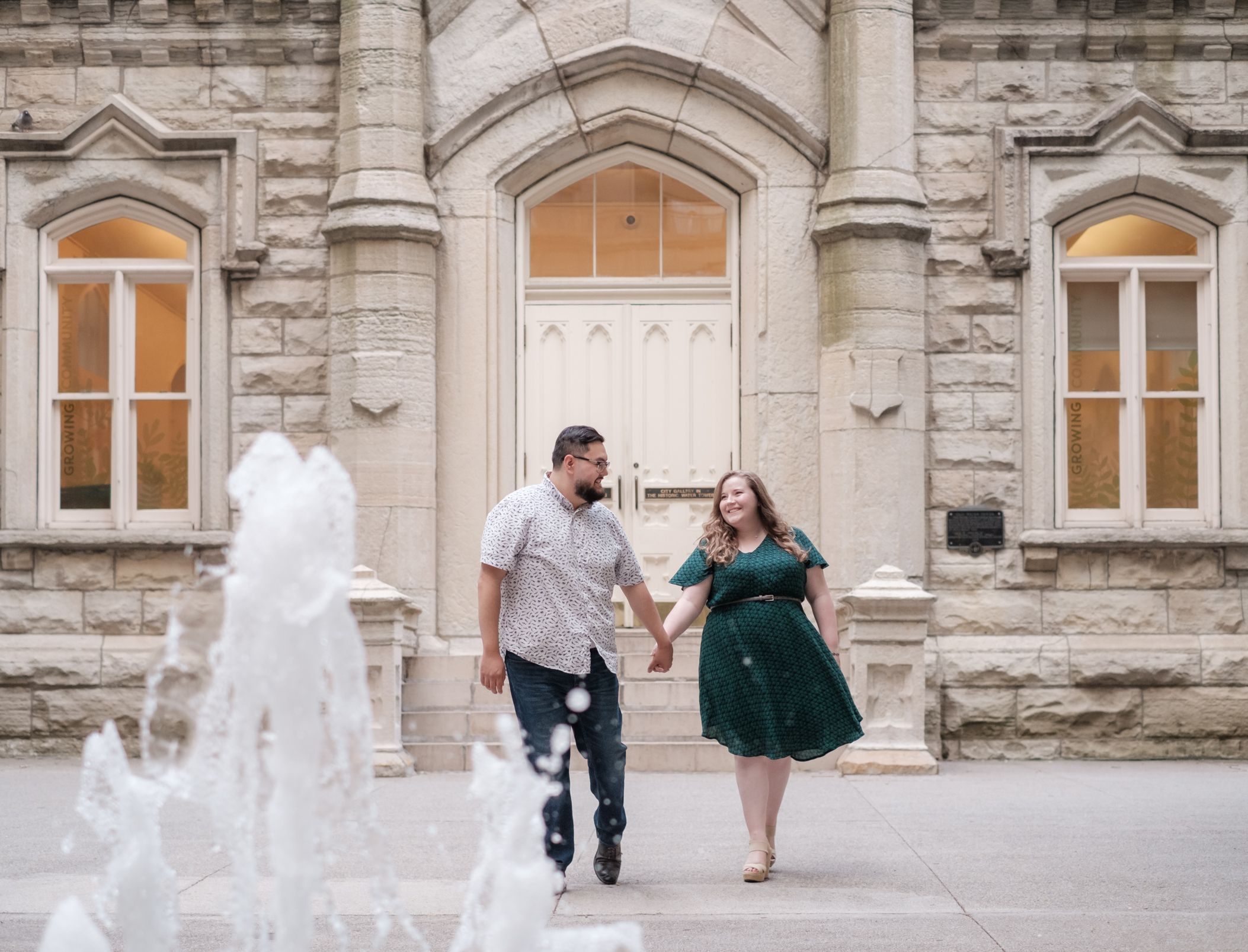 A couple joyfully runs and dances together in front of a historic stone building with arched windows and a fountain.