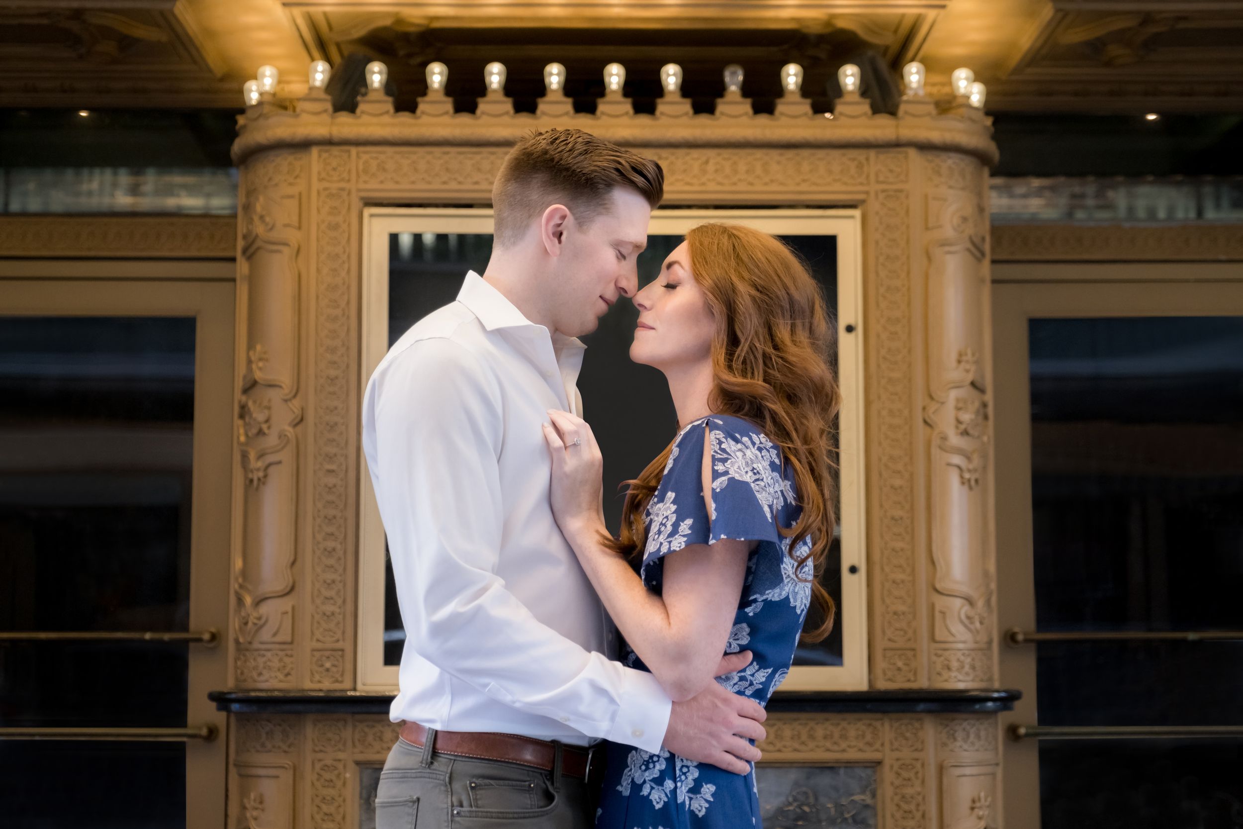 A romantic couple embraces in front of an ornate gold-framed window in an elegant historic building.