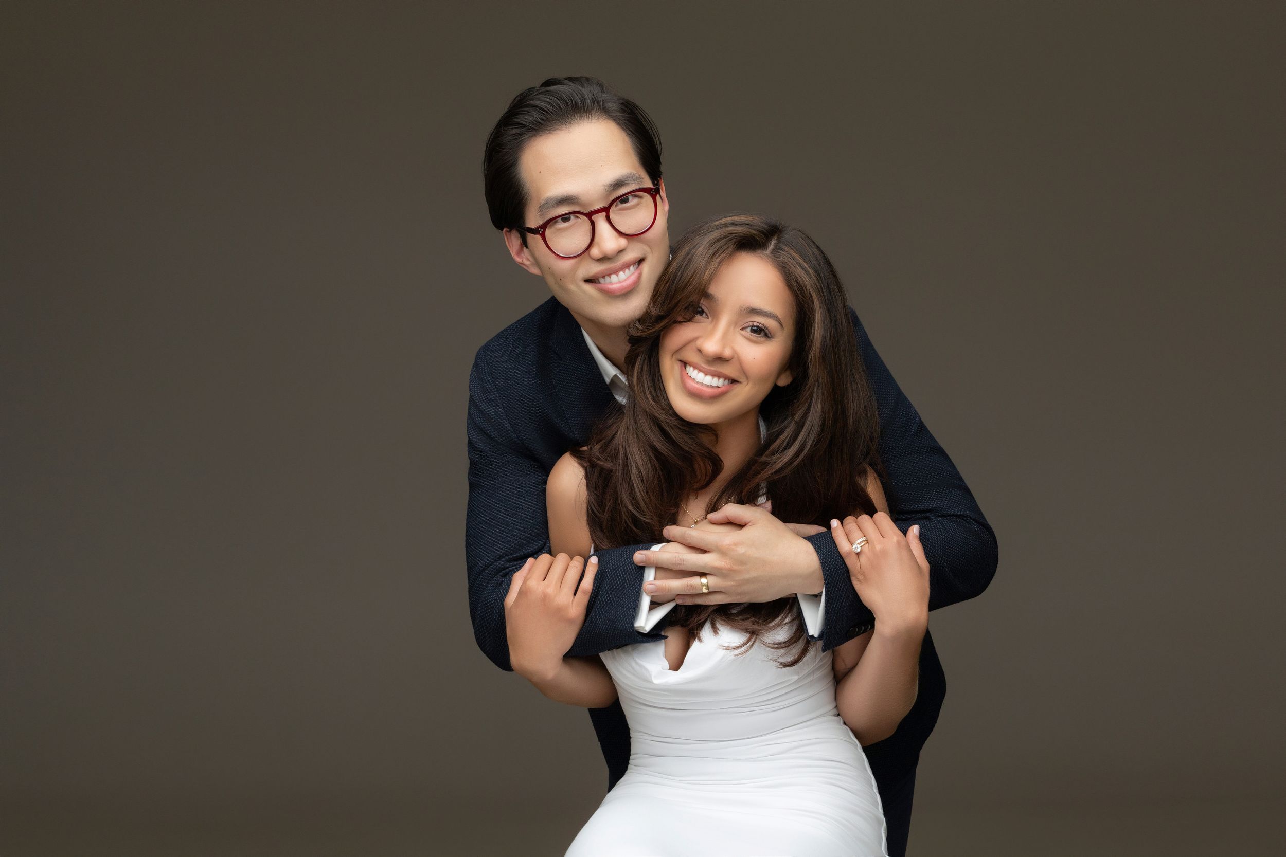 A couple in formal attire embrace and smile warmly against a dark background during a wedding photoshoot.