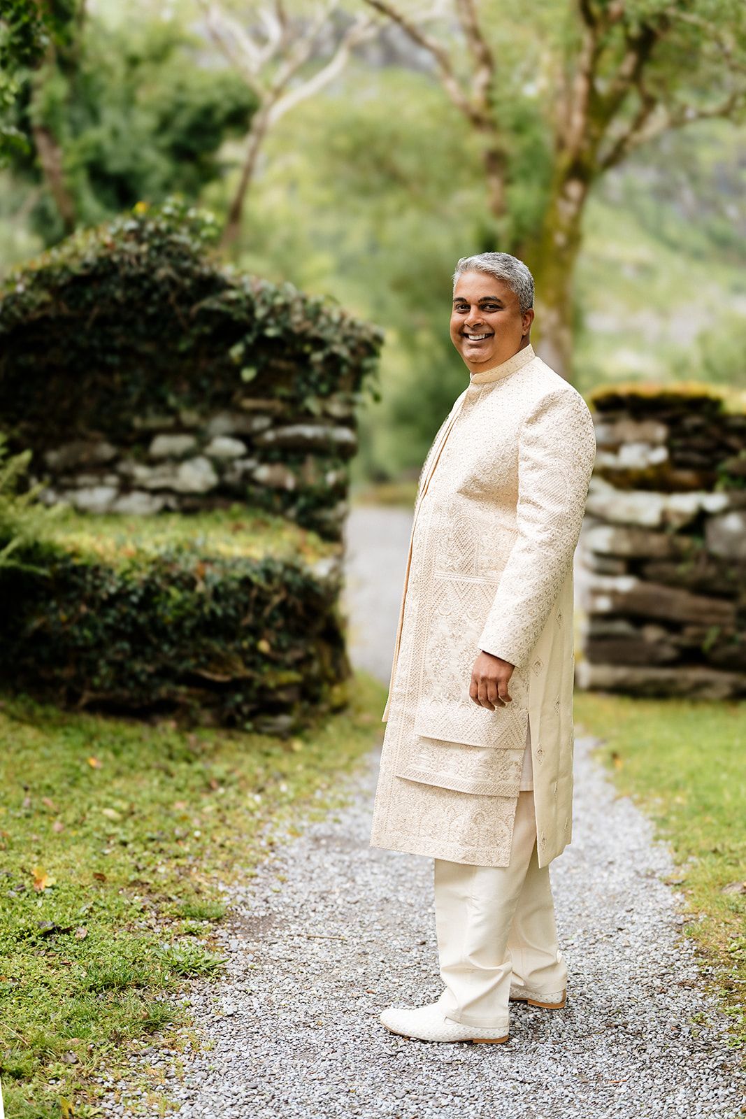 Someone wearing an elegant cream-colored traditional Indian kurta stands on a gravel path surrounded by lush greenery.