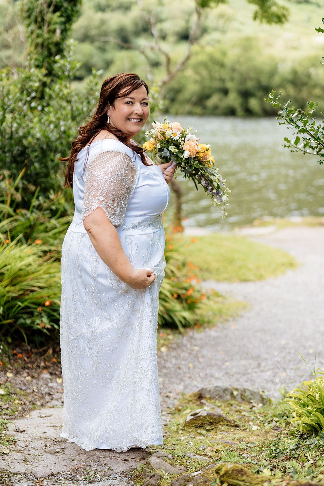 Wedding photos showing person in white lace dress holding sunflower bouquet on path by scenic lake.