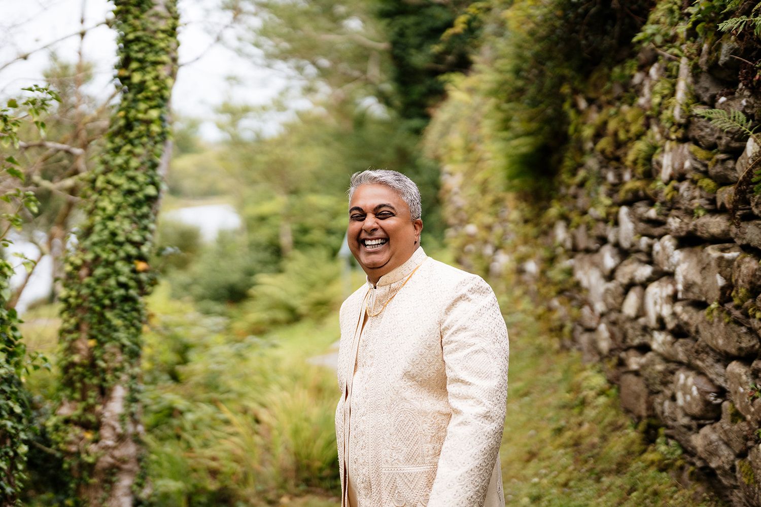 A smiling person in a cream-colored jacket stands beside an ivy-covered stone wall in a lush garden setting.