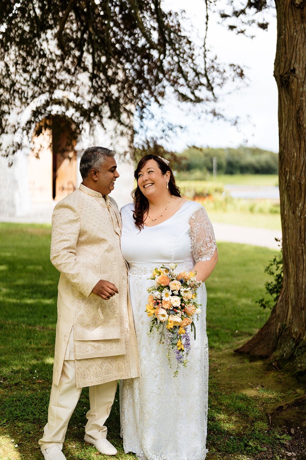 A couple in cream-colored wedding attire share intimate moments outdoors under tall trees in a natural setting.