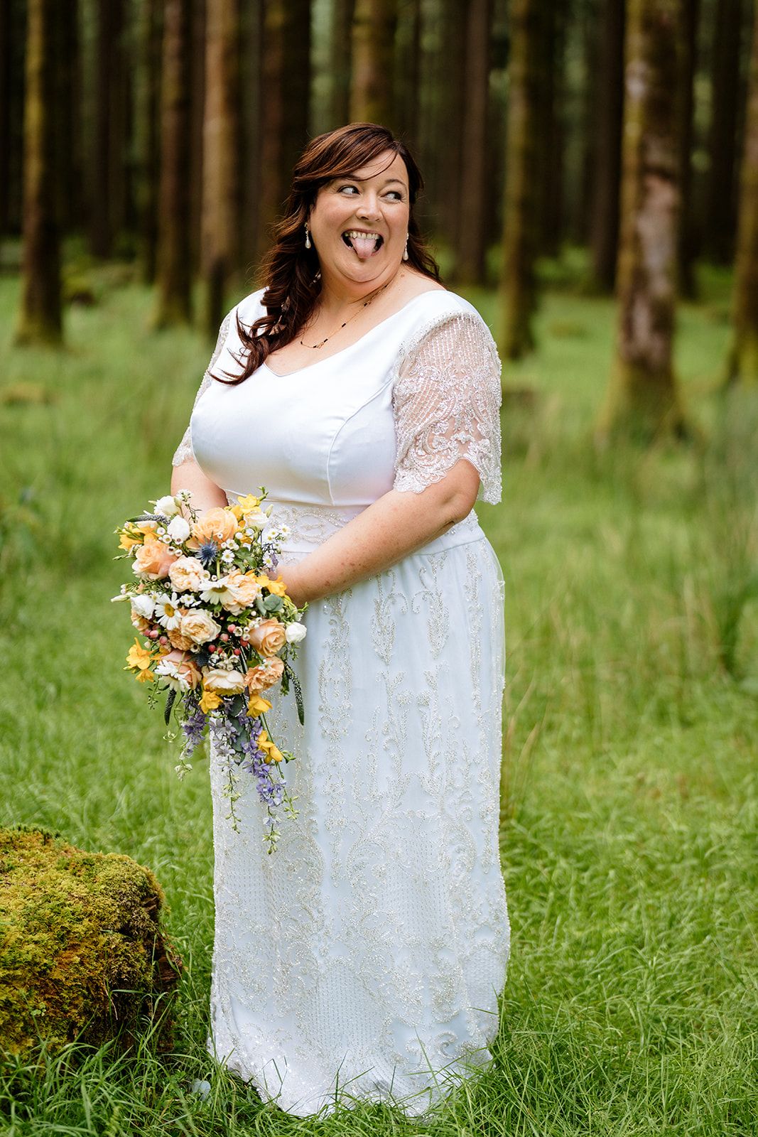 A bride in a white lace wedding dress stands in a forest setting holding a colorful yellow and white floral bouquet.