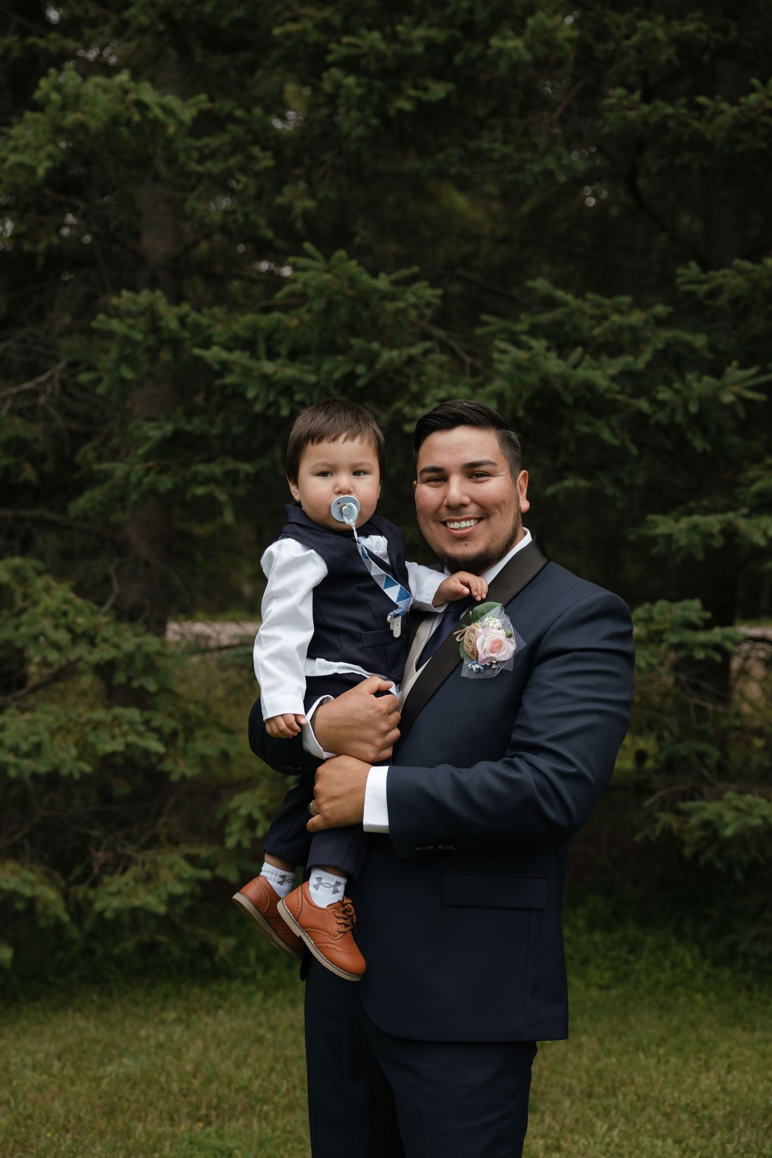 A tender moment captured outdoors between two people in formal attire standing among trees and greenery.