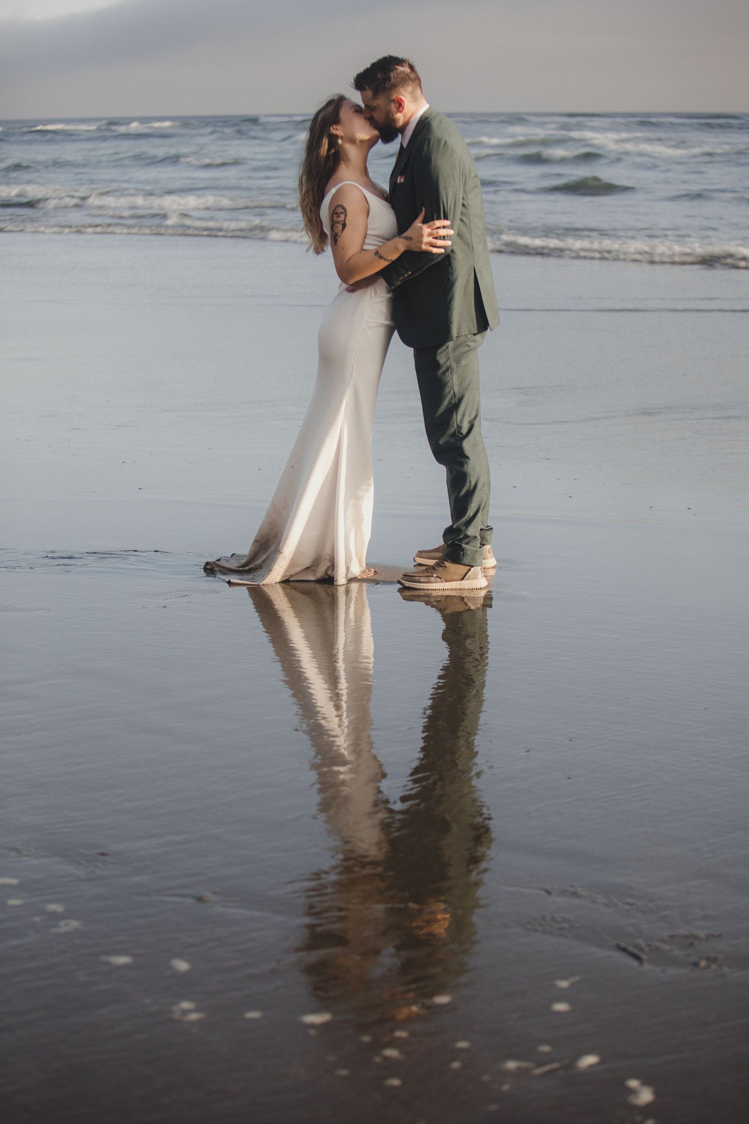 A romantic couple shares an intimate moment on a reflective beach at sunset in formal attire.
