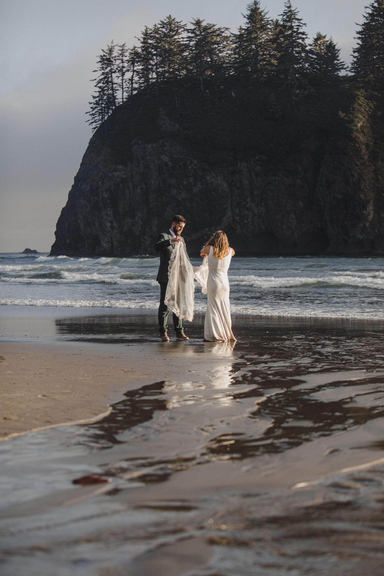 Two people in white dresses stand together on a misty beach with a dramatic sea cliff in the background.