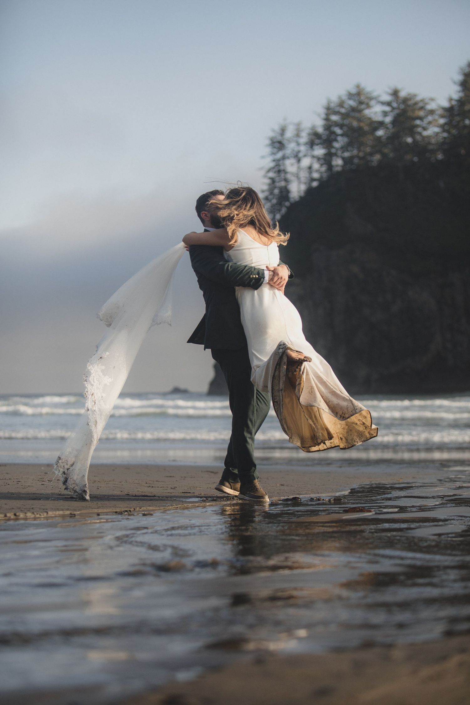 Wedding couple dances on a misty beach with waves and evergreen forest in the background during golden hour.