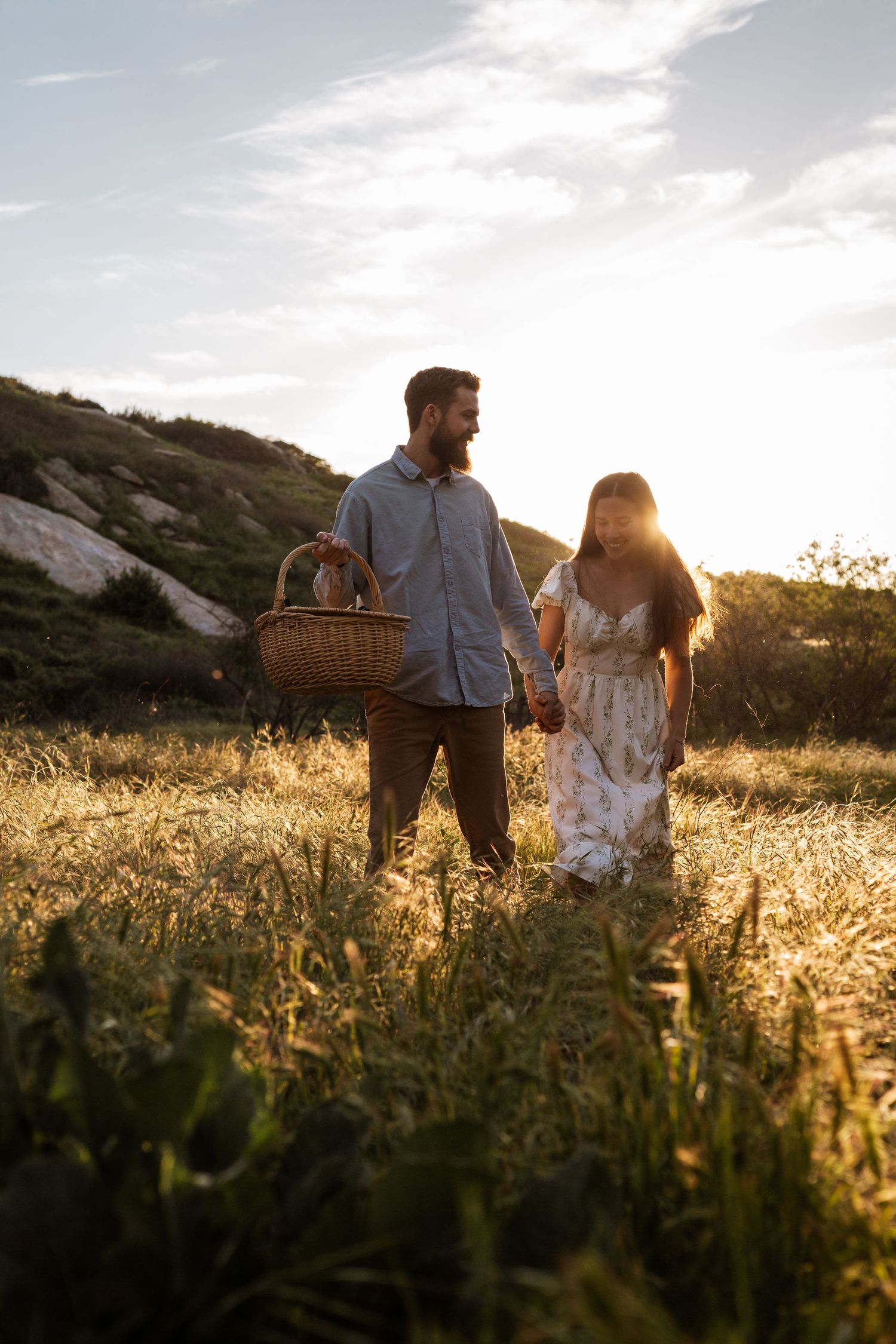 Couple walking through golden field at sunset carrying picnic basket.