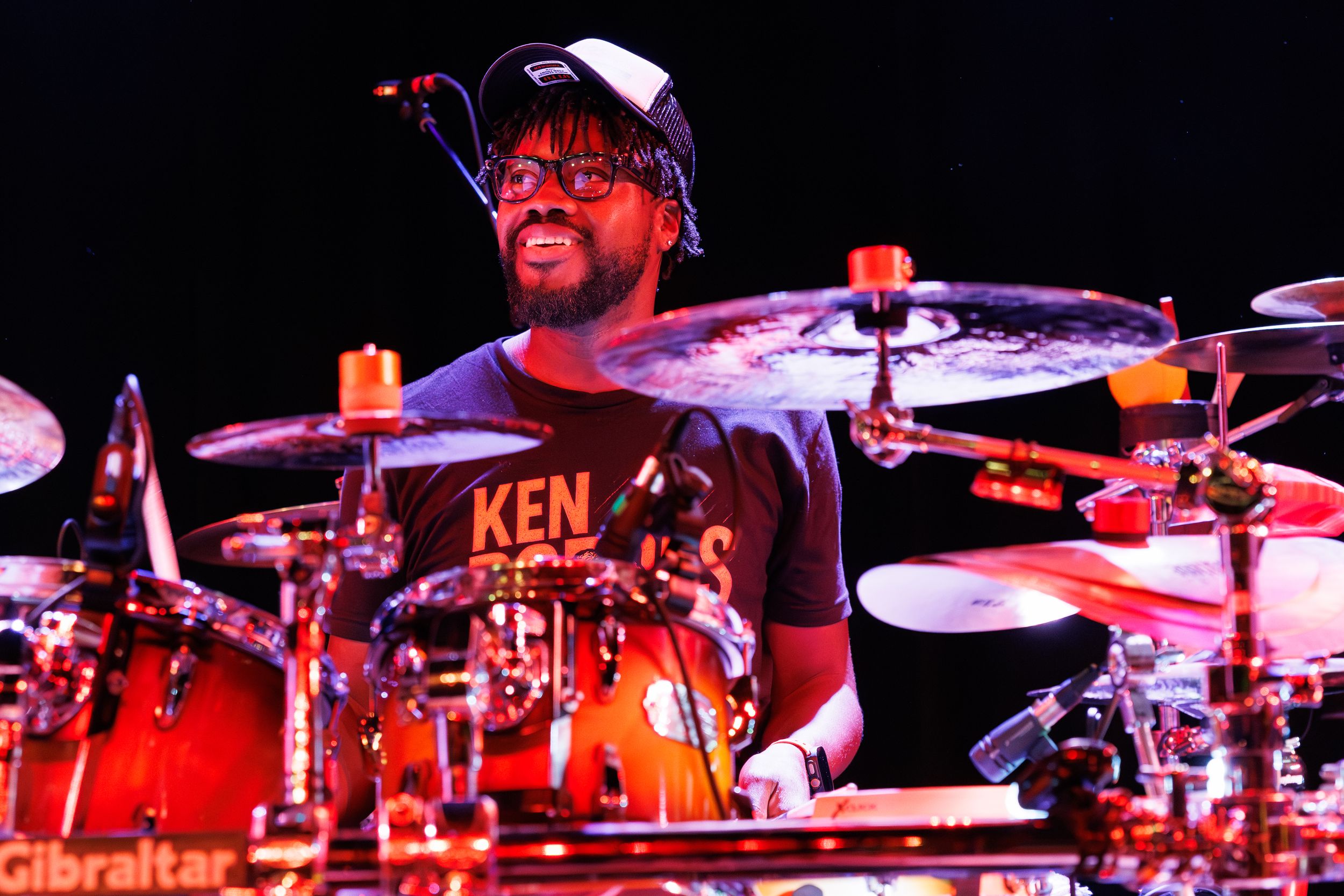 A drummer performs behind a red drum kit illuminated by dramatic red stage lighting during a live performance.