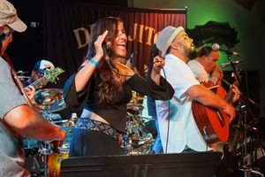 Musicians perform energetically on stage with colorful lighting and instruments at a live concert event.