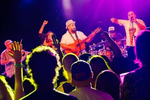 Energetic band performs on stage while silhouetted crowd watches from below with purple and blue stage lighting.