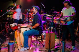 Band members play percussion instruments and drums on stage with colorful red and purple lighting effects.