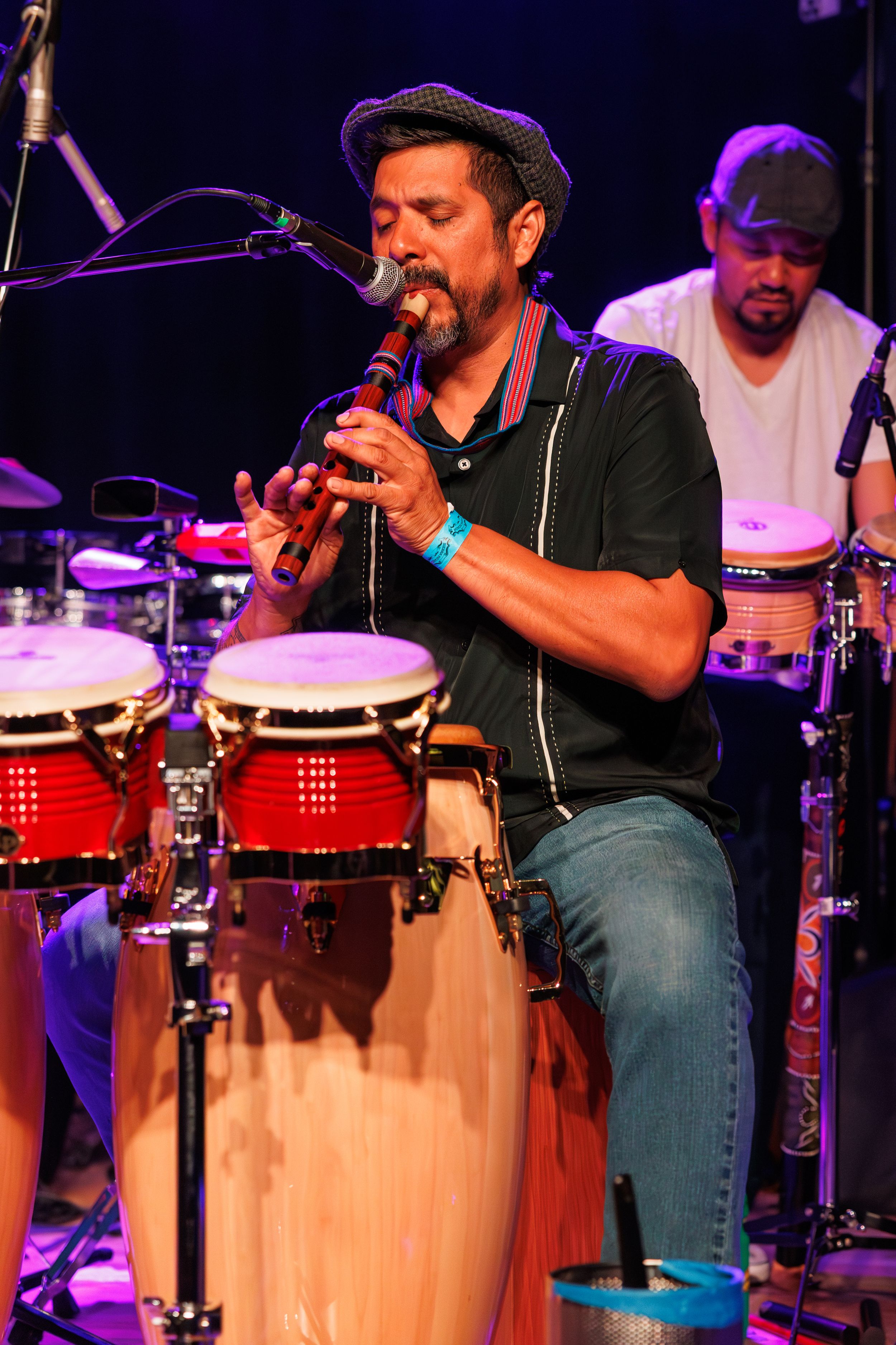 A percussionist performs on congas during a live concert with dramatic purple stage lighting.