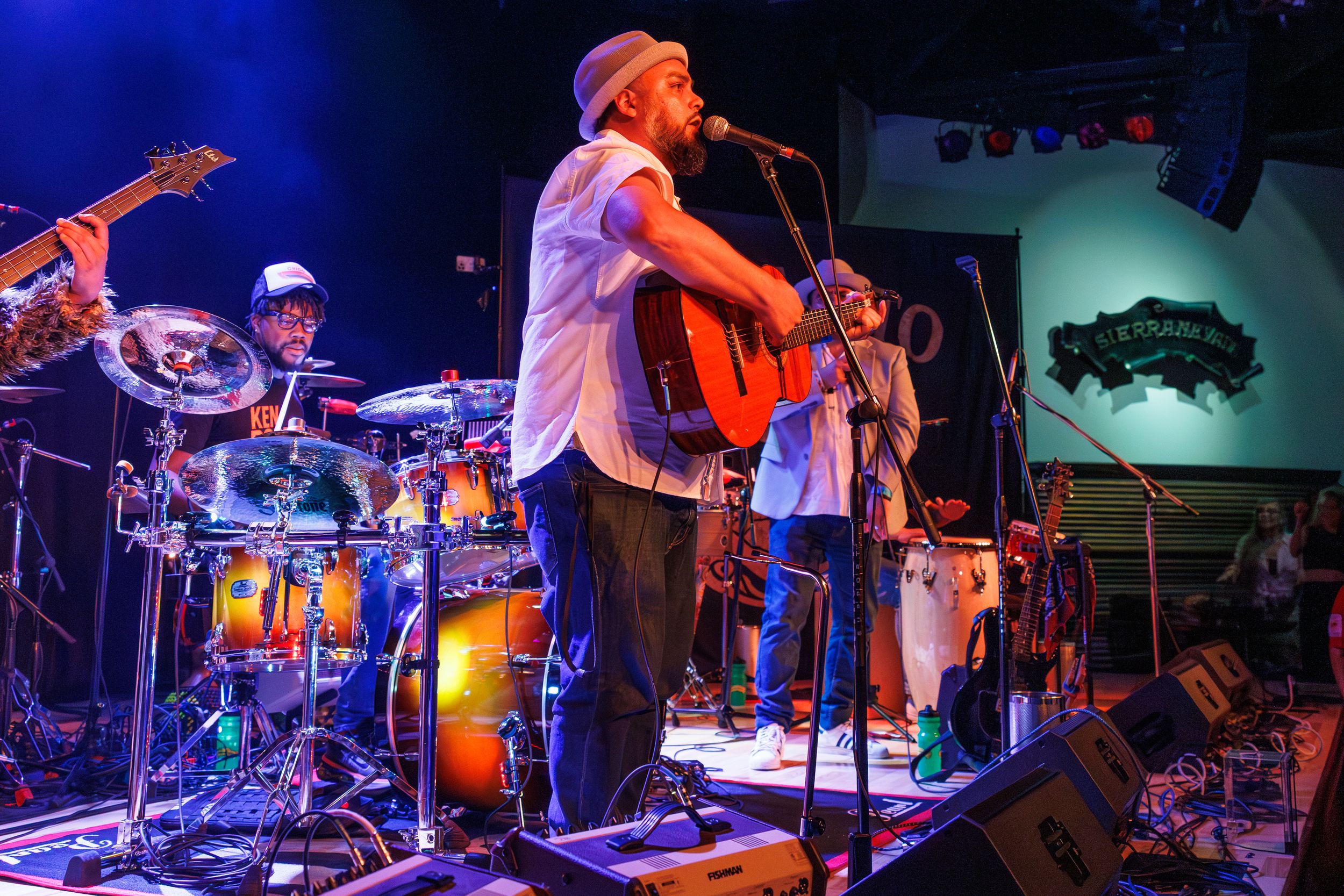 Musicians perform on stage with acoustic guitar and drum kit under blue stage lighting at live concert venue.
