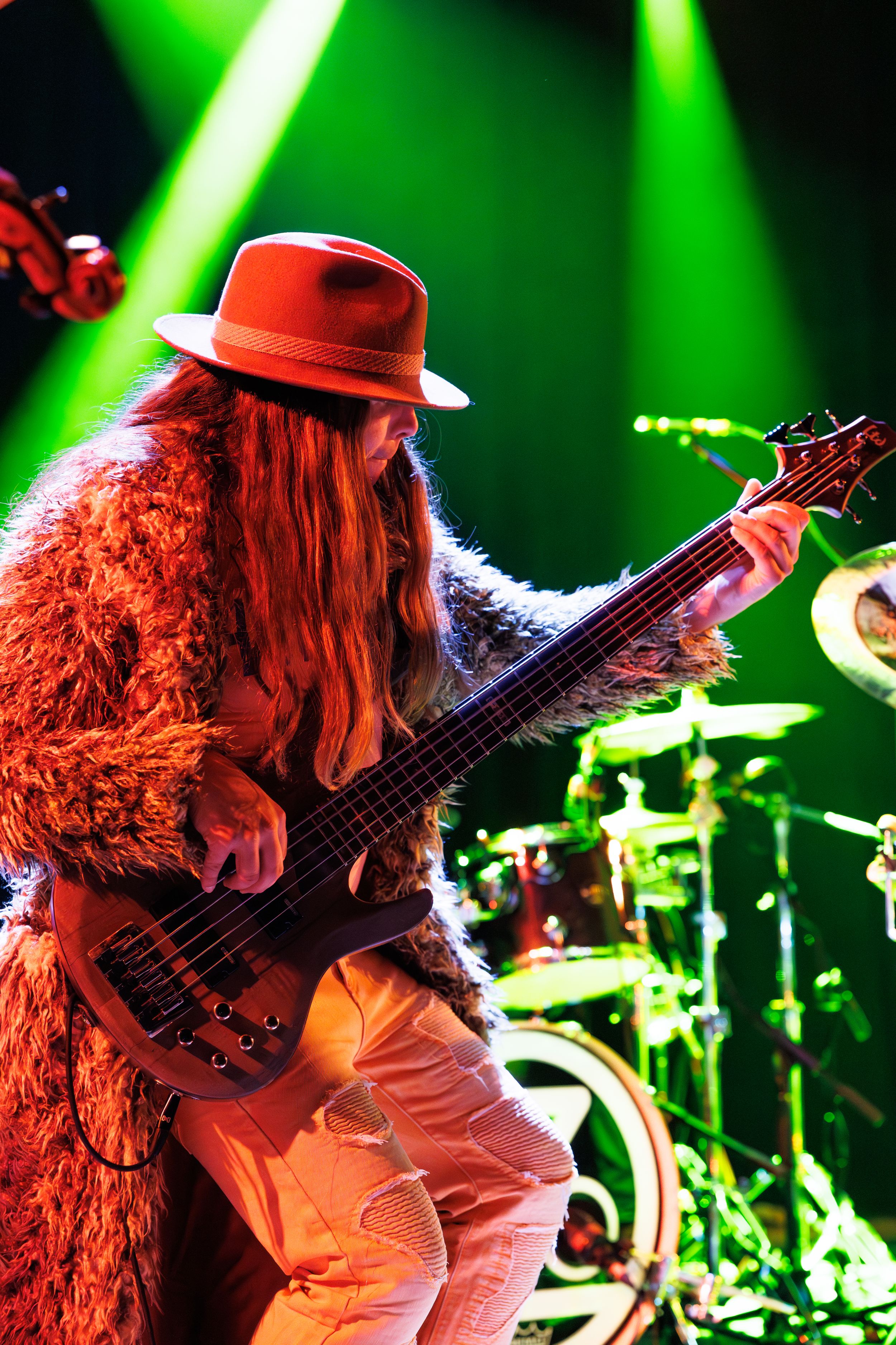 Musician performing with long hair and a hat plays bass guitar under green stage lighting.