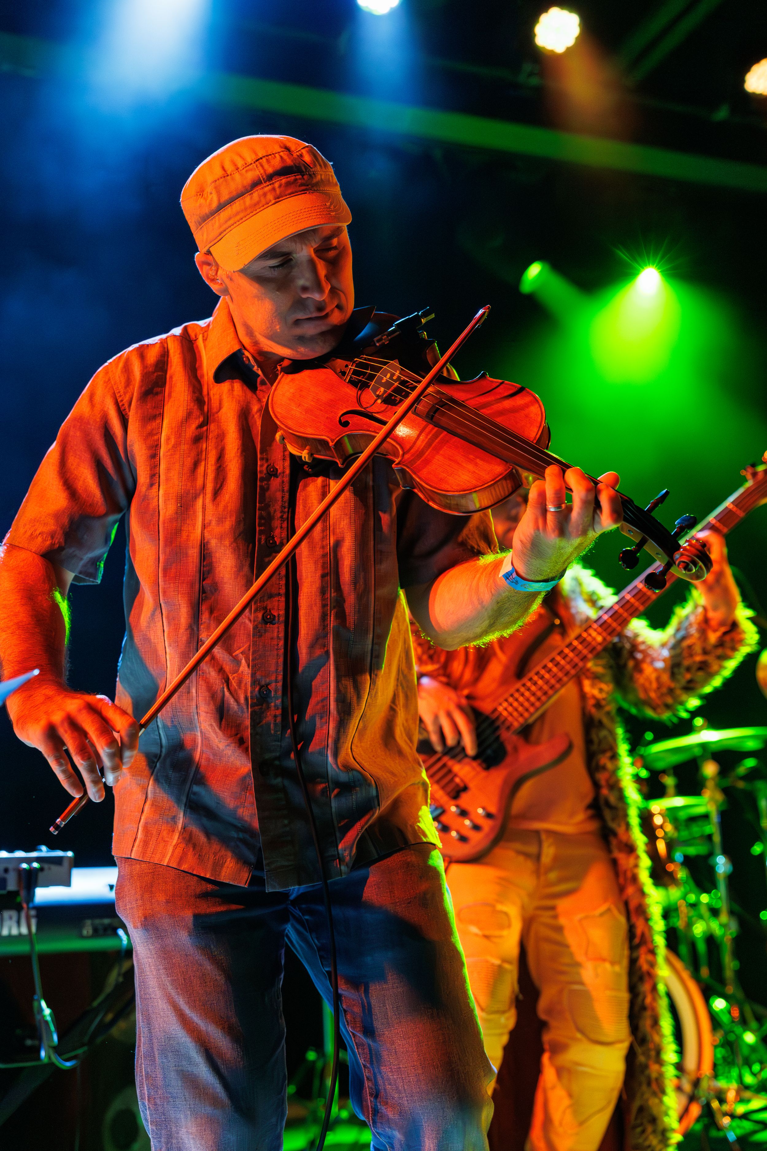 Musician plays electric violin on stage with colorful green and blue lighting during a live performance with band.