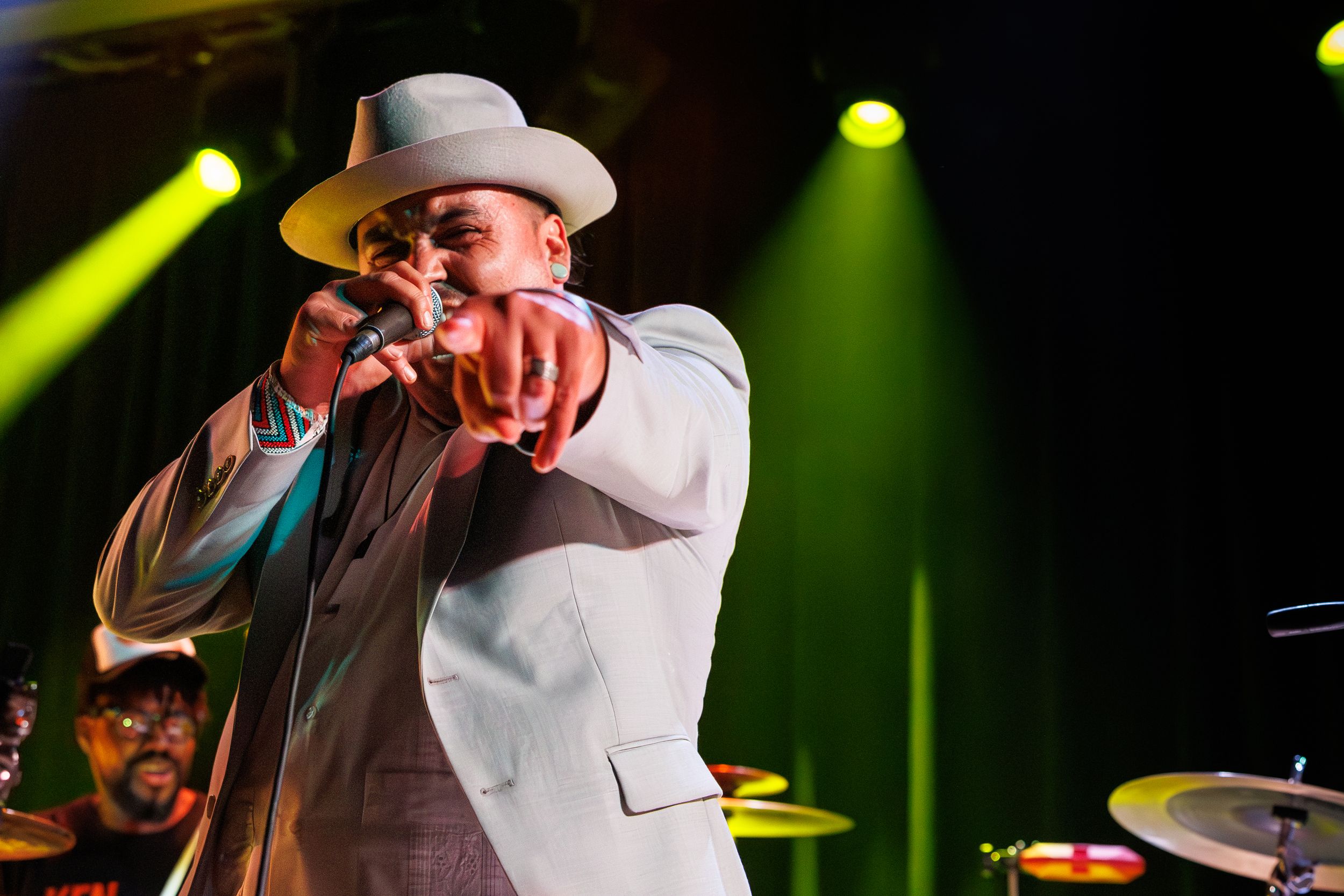 A performer in a white suit and hat gestures energetically on stage with colorful spotlights in the background.
