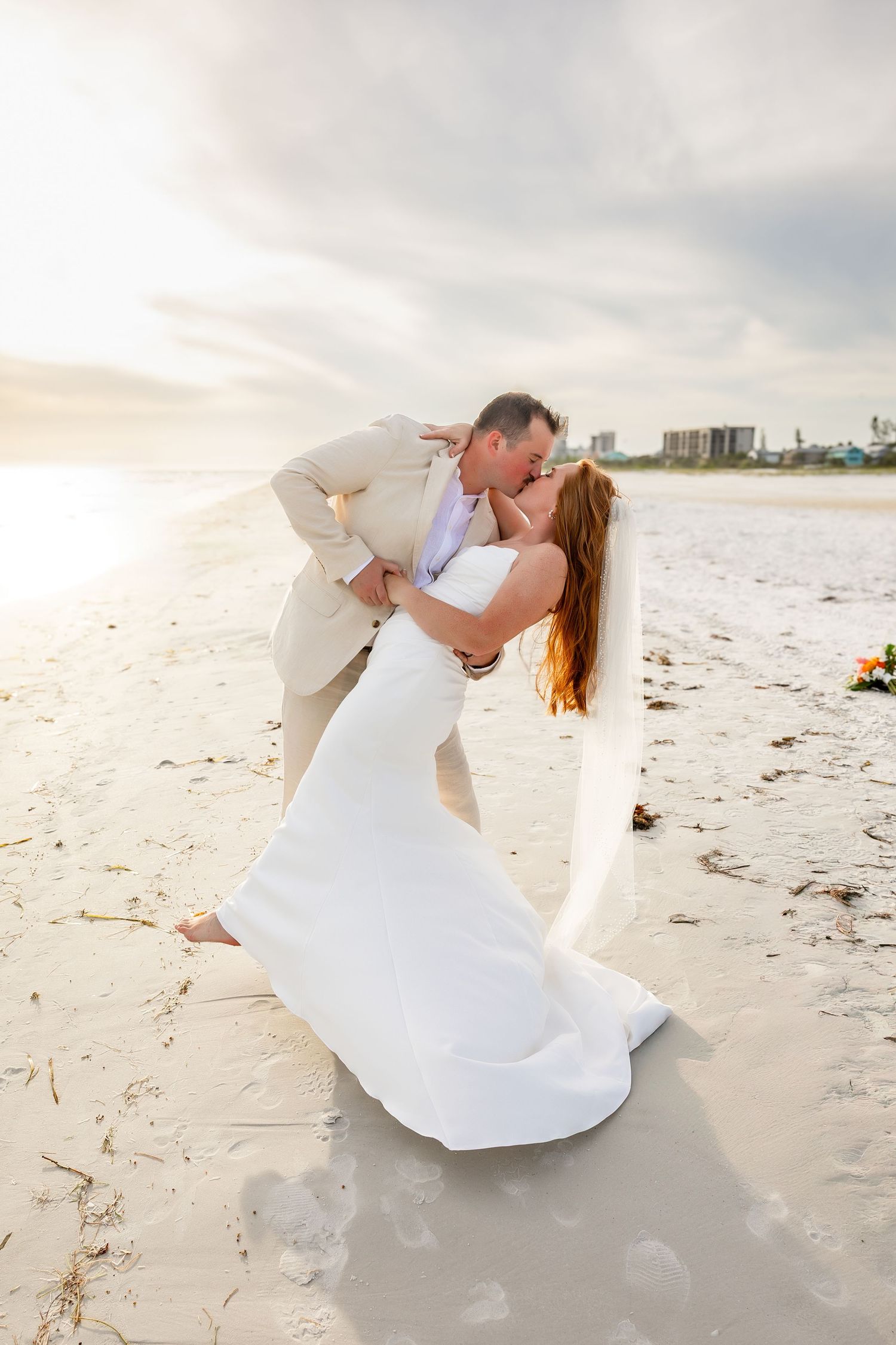 Bride and groom sharing romantic dip kiss on white sand beach at sunset during wedding ceremony.