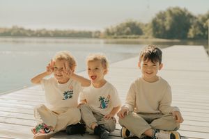 Three children in matching beige sweaters sit together on a wooden dock by a serene lake during sunset.
