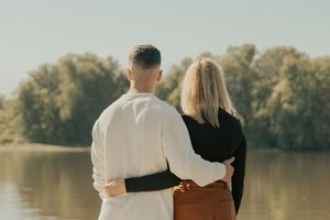 A couple stands together by a lake's edge during an engagement photoshoot in a tranquil outdoor setting.