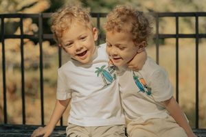 Two children in white t-shirts share a heartwarming laugh together while sitting outdoors by a black metal fence.