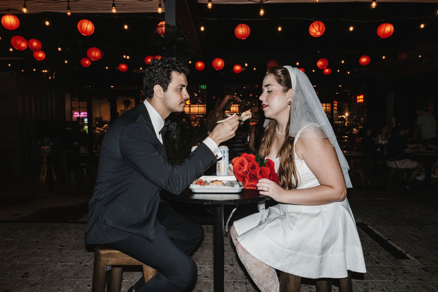 A couple shares an intimate moment at a dimly lit restaurant table decorated with red lanterns overhead.