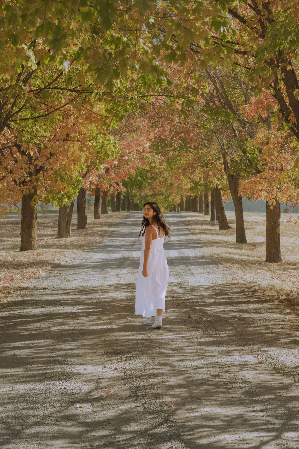 Senior girl wearing a white tank dress walking away and looking back at camera smiling during fall with the leaves changing color.