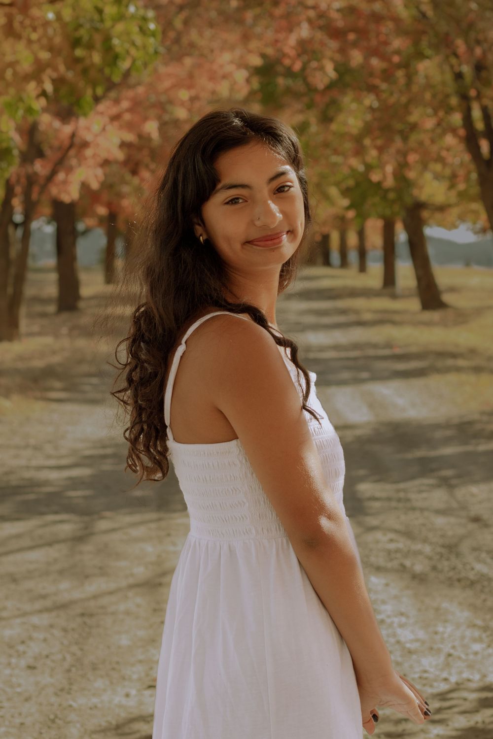 High school graduate smiling in a white dress in the center of a autumnal tree lined driveway.