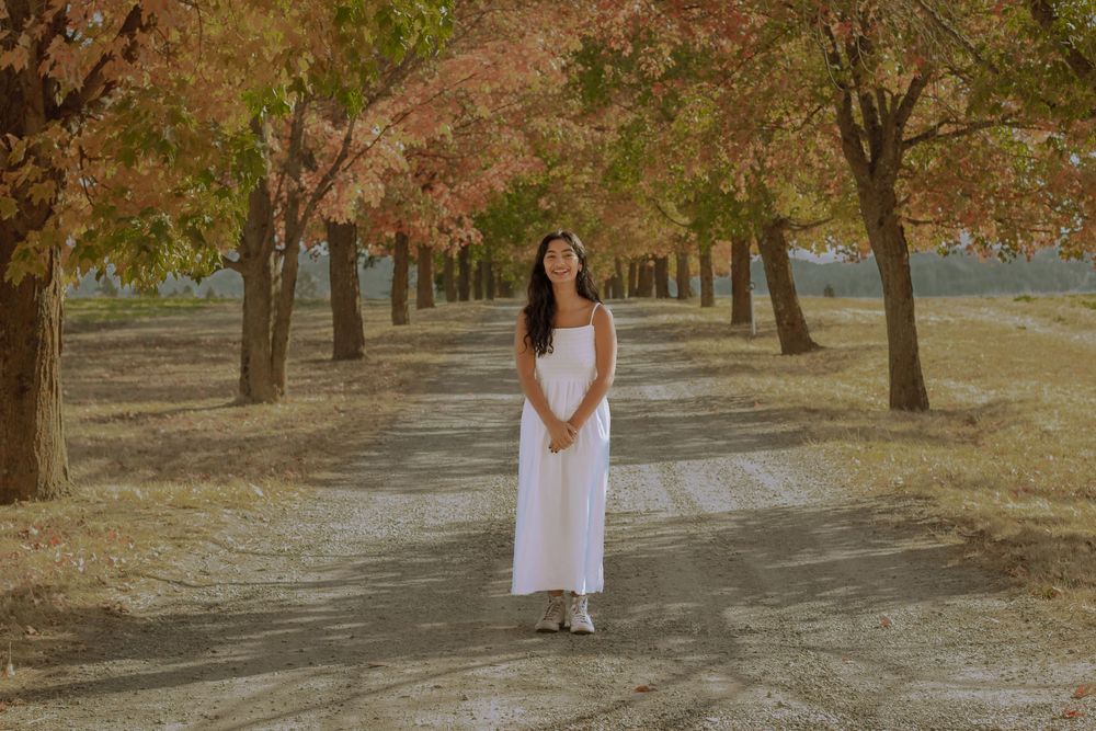Woman in long white dress posed in front of tree lined driveway during fall with colorful scenery.