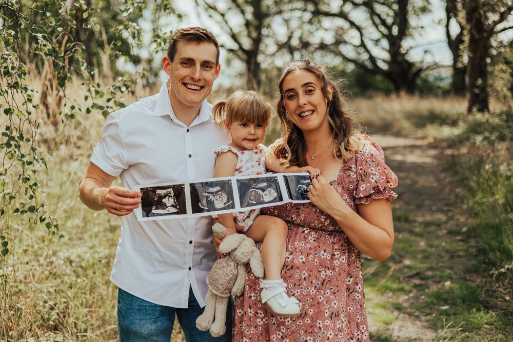 Happy couple in a pink floral dress and white shirt hold ultrasound photos and hug in an outdoor nature setting.