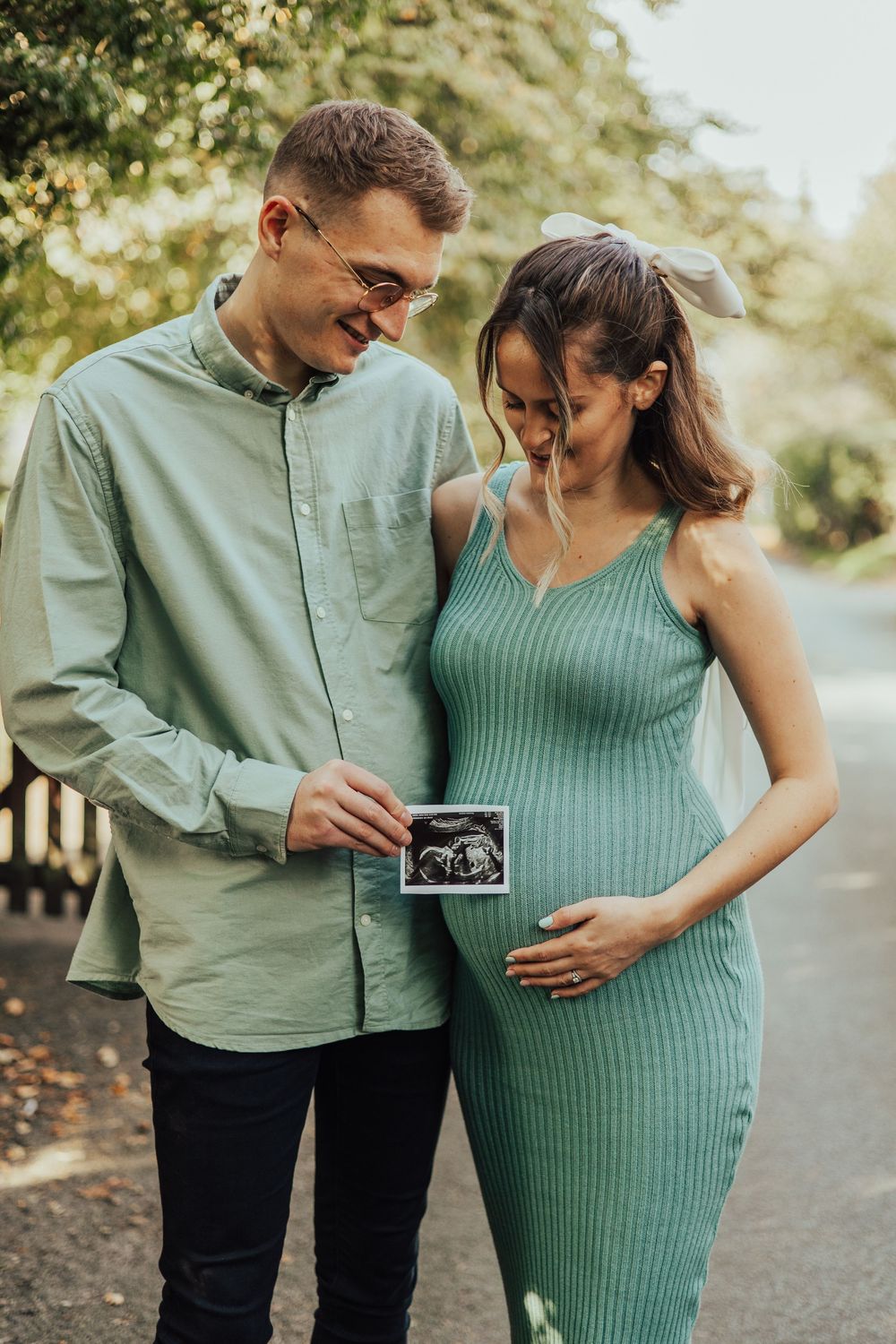 Expecting couple in matching green outfits holds ultrasound photo while smiling on outdoor path.