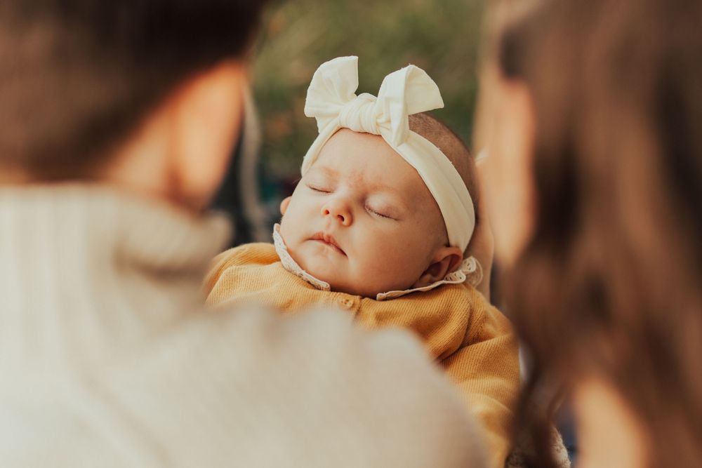 A sleeping infant wearing a white bow headband and yellow outfit is held between two people in soft lighting.