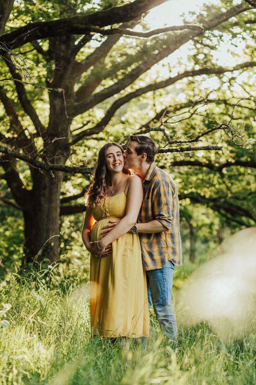 A couple shares intimate romantic moments in a sun-dappled meadow beneath tall trees during a summer afternoon.