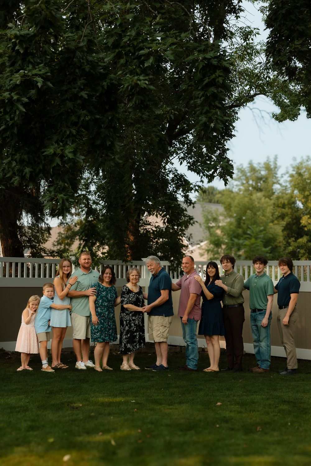 A group of people standing together on a lawn at dusk with trees and a white fence in the background.