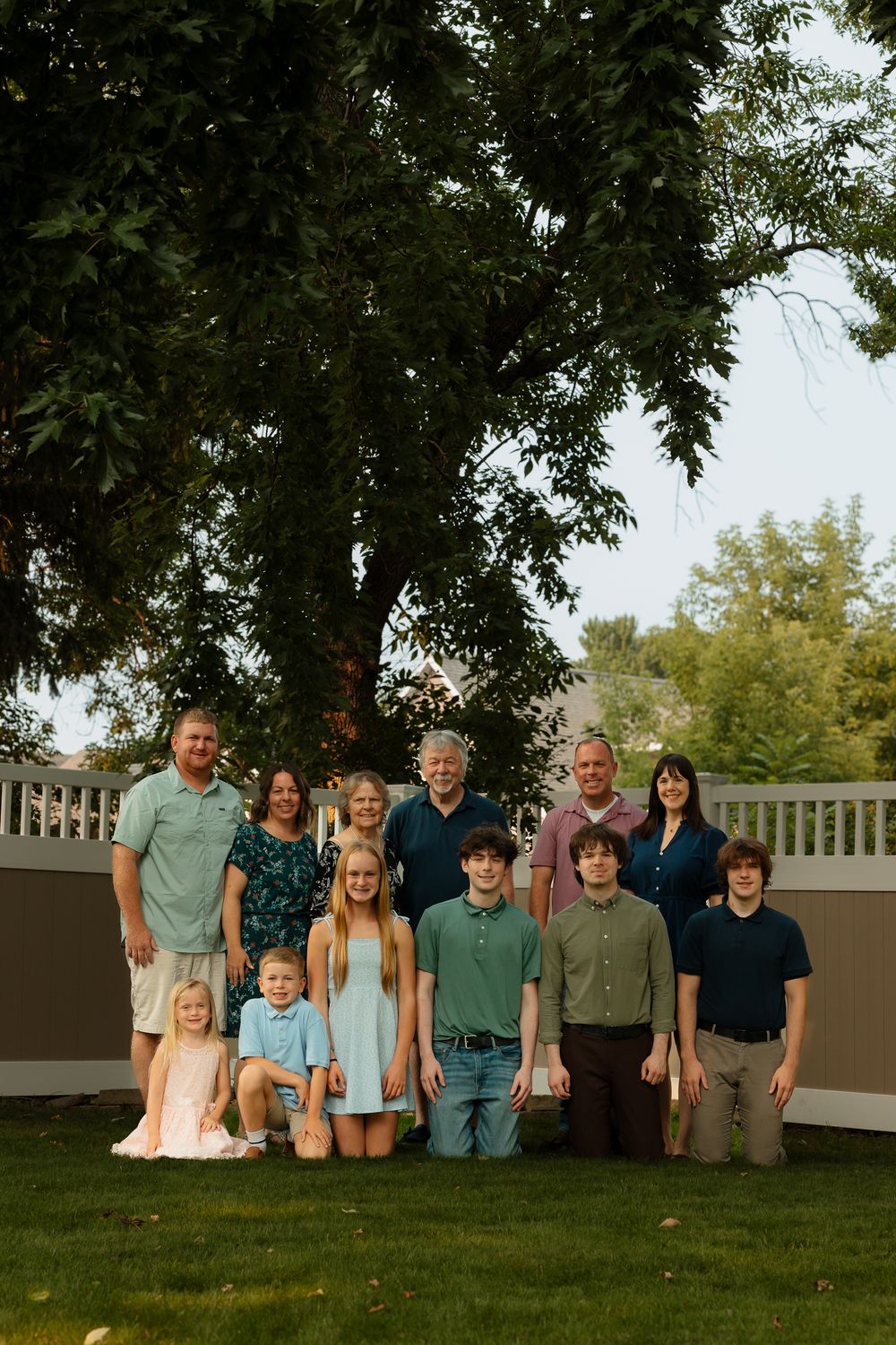 A group of people pose together outdoors on a grassy lawn under large trees during a casual photo session.