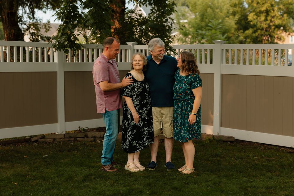 People gather and converse on a lawn area in front of a white picket fence during golden hour lighting.
